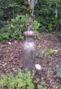 Brass tone urn with a sparkler hummingbird inside. The sprinkler head is barely visible above the flower.